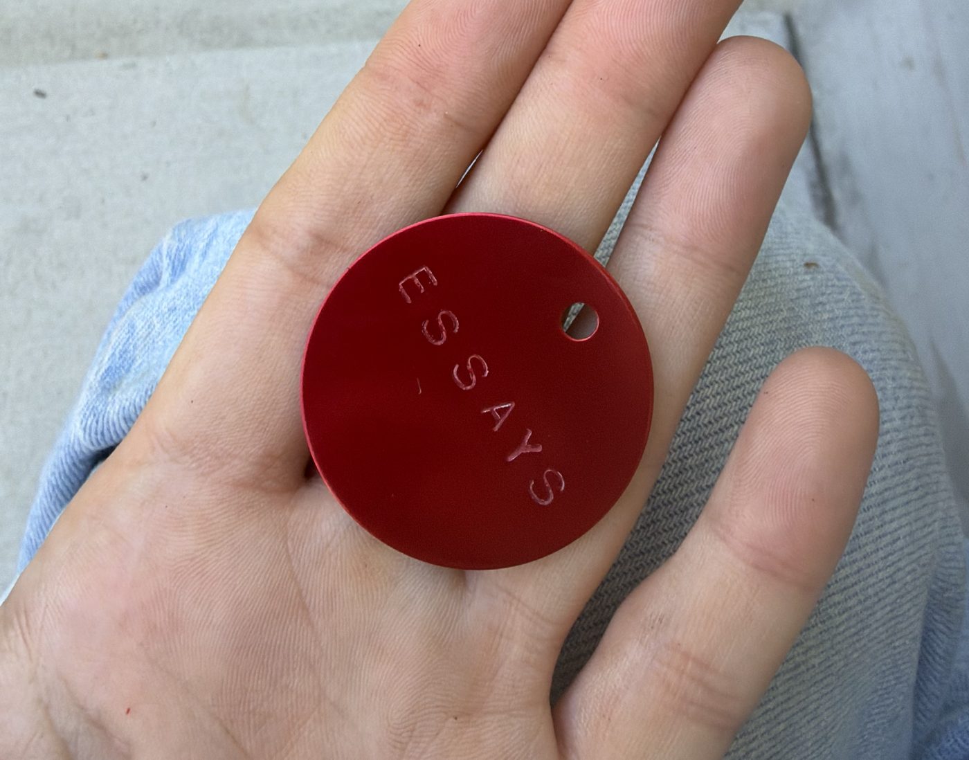 A red anodized aluminum medallion stamped with the word ESSAYS, held in a hand